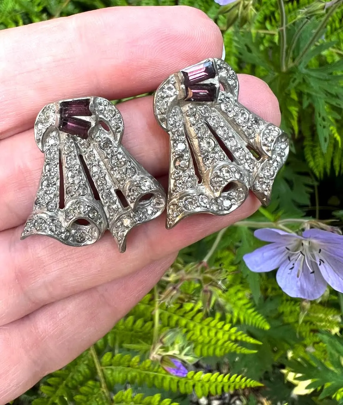 Two Art Deco dress clips held in a hand outdoors with ferns and purple flower