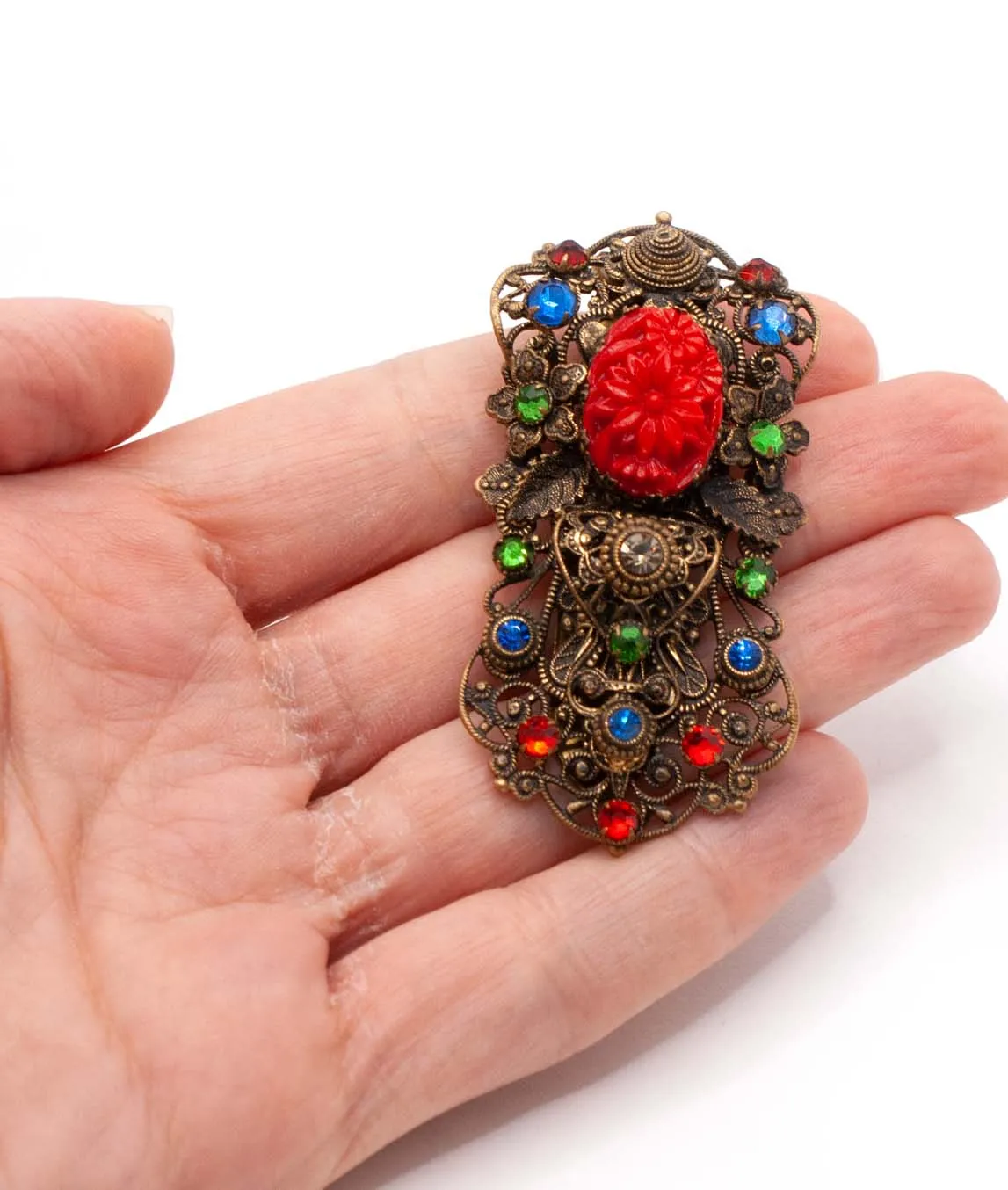 1930s Czech pierced metal dress clip decorated with red green and blue crystals and red pressed glass held in hand