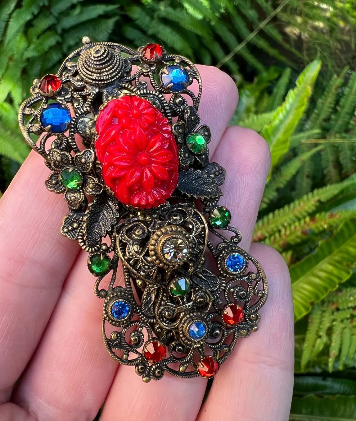 1930s Czech pierced metal dress clip decorated with red green and blue crystals and red pressed glass in hand on greenery